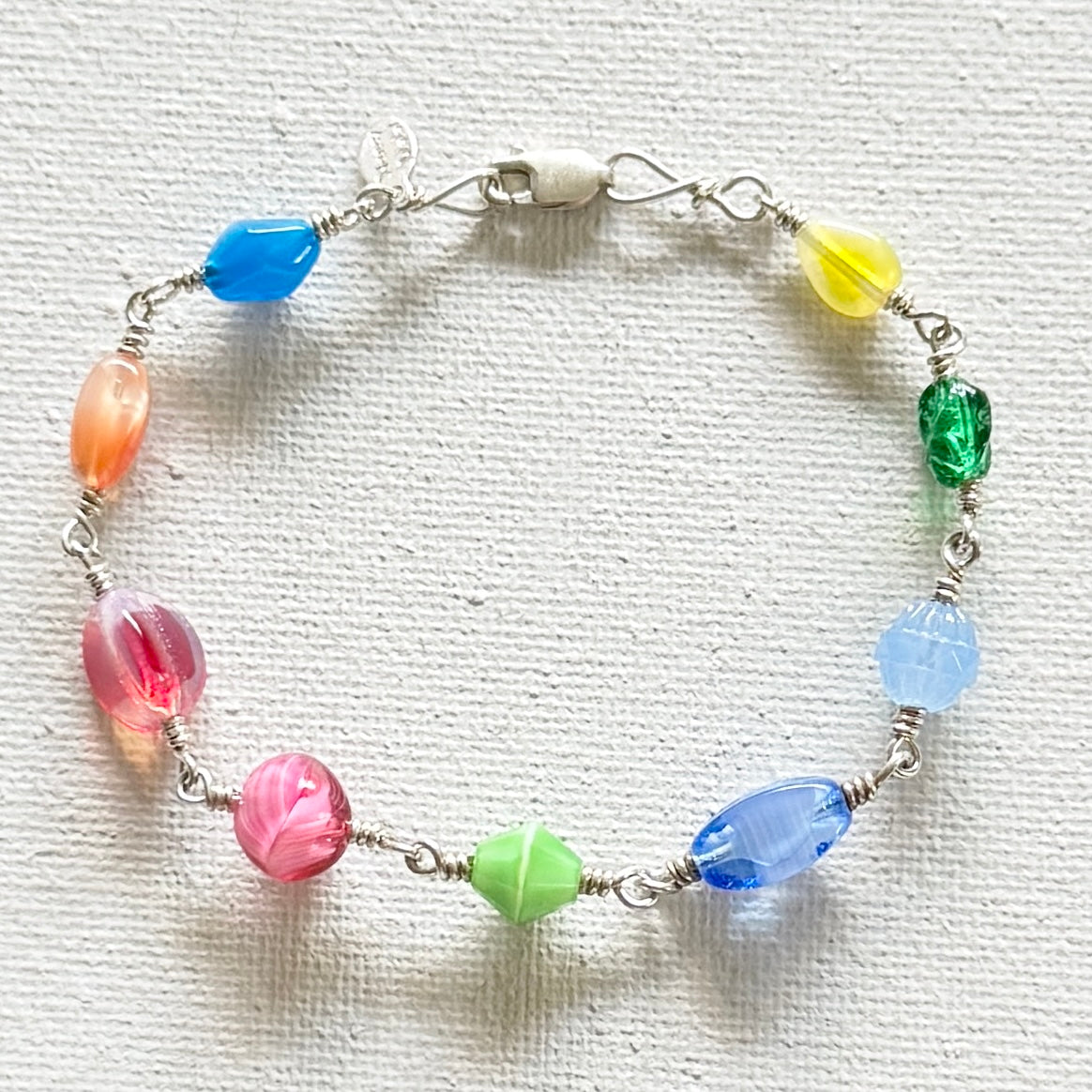 A handmade wire-wrapped beaded bracelet featuring nine colorful, faceted glass beads in shades of blue, yellow, green, pink, and light blue, arranged in a circle on a textured white fabric background.