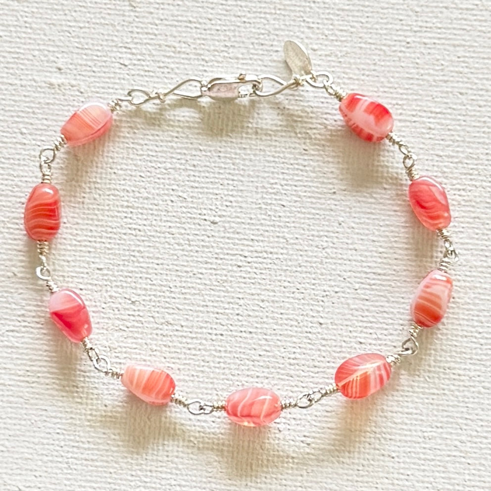 A silver hand linked bracelet featuring multiple red and white marbled beads, displayed on a textured off-white fabric background.