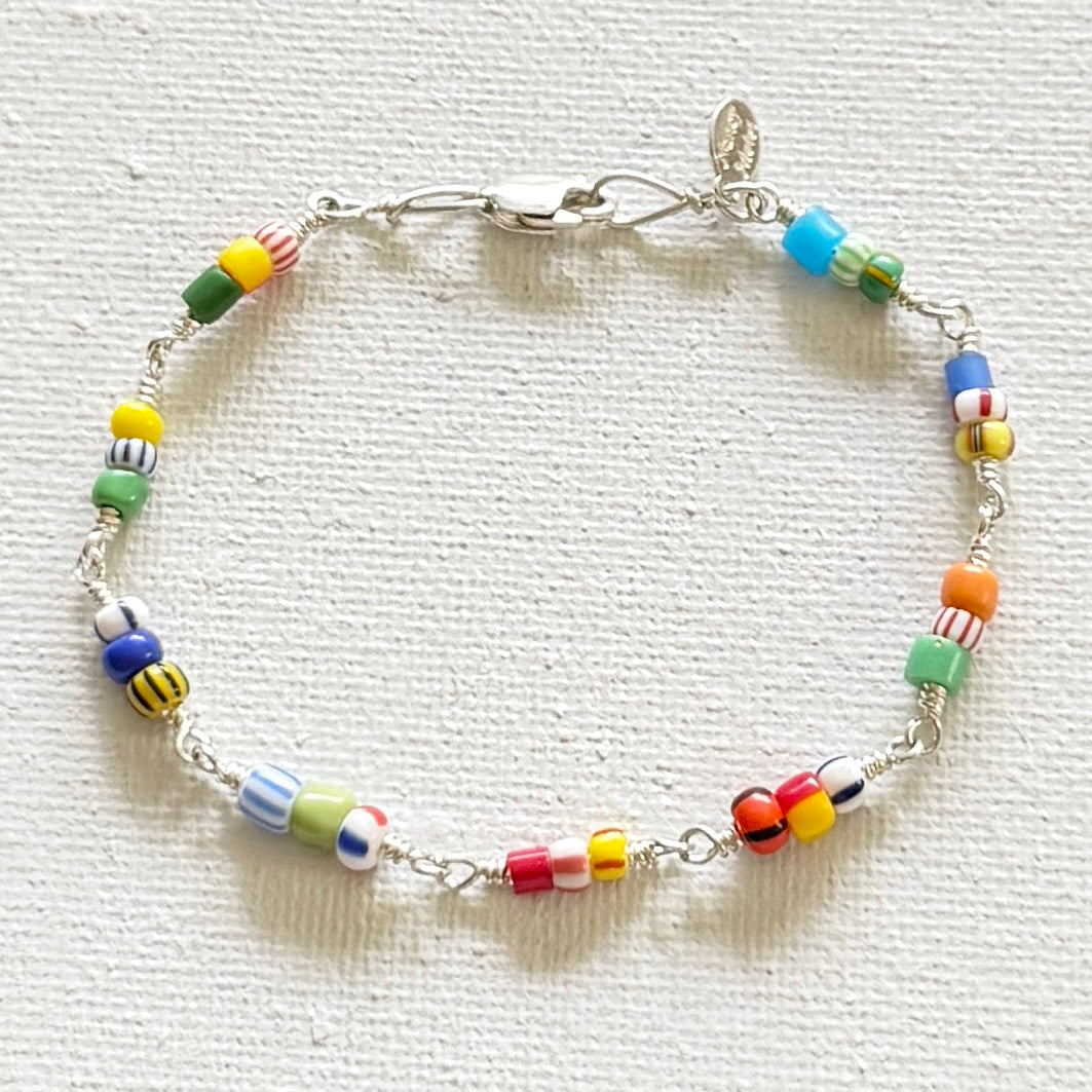 A silver linked bracelet adorned with colorful, patterned cylindrical beads in yellow, green, blue, orange, red, and white, displayed on a textured off-white fabric background.