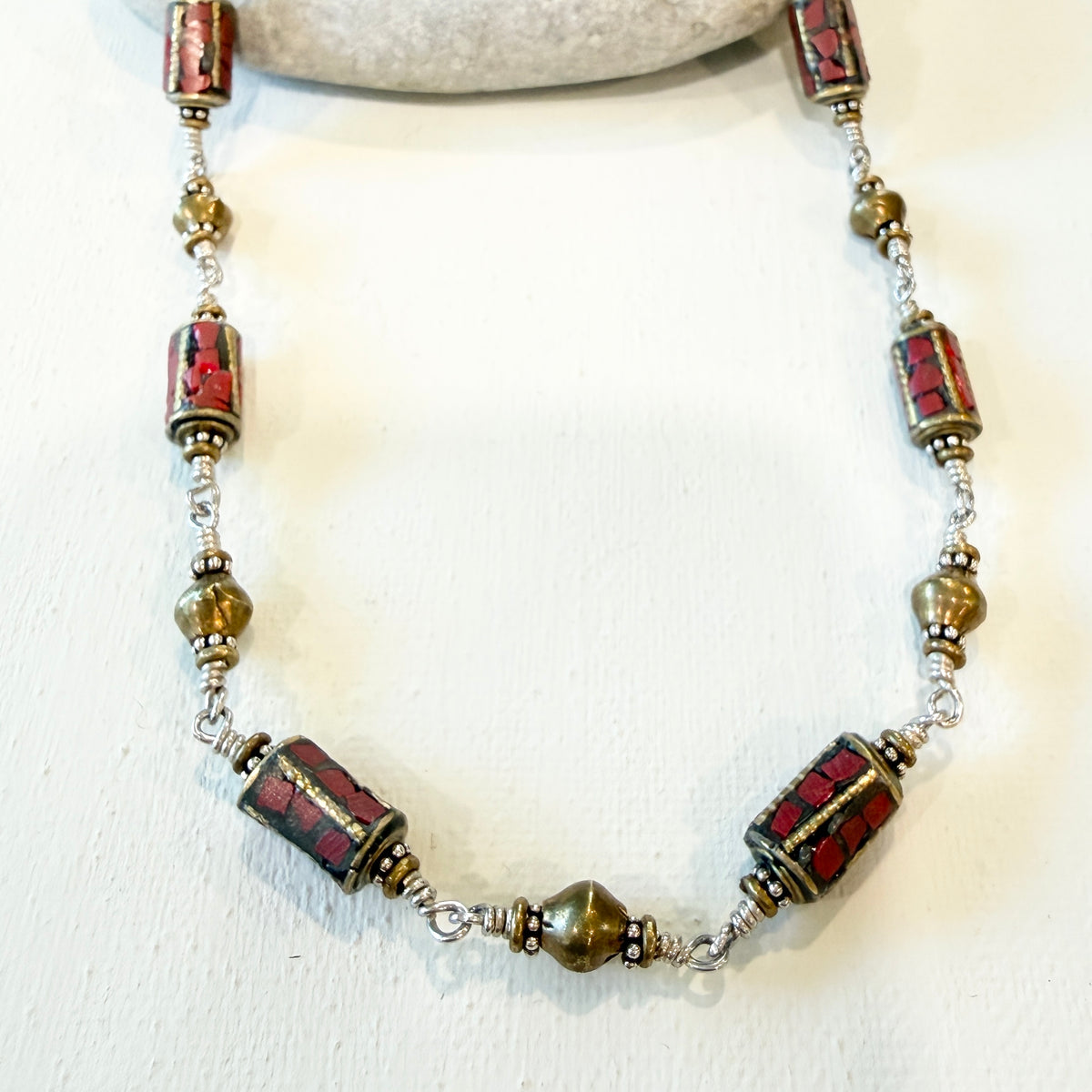 A handmade beaded necklace featuring cylindrical red and black patterned beads, small metallic bronze beads, and silver-toned wire spacers, displayed on a white surface.
