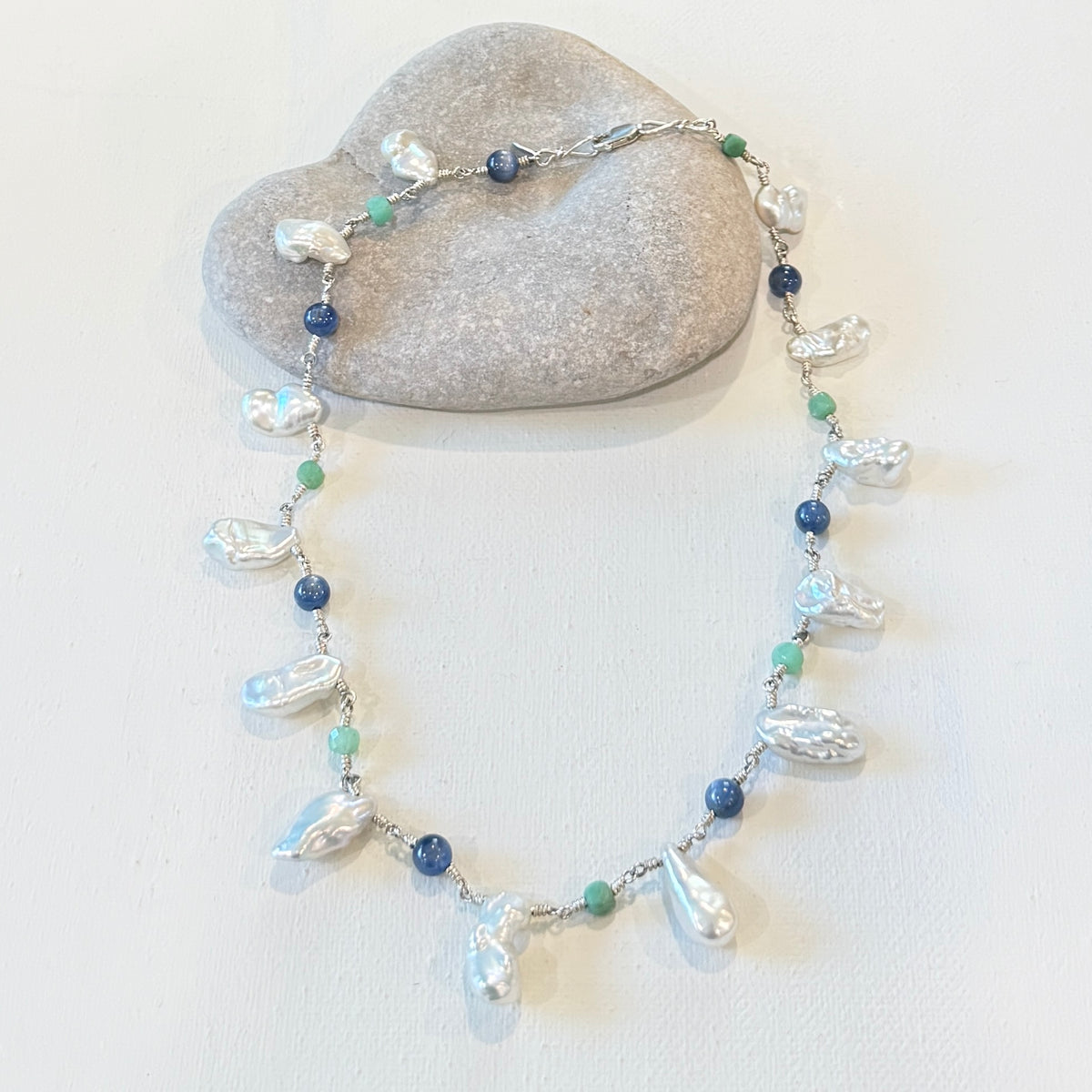 A beaded necklace with baroque pearls, blue beads, and green beads, displayed on a small gray stone against a white background.