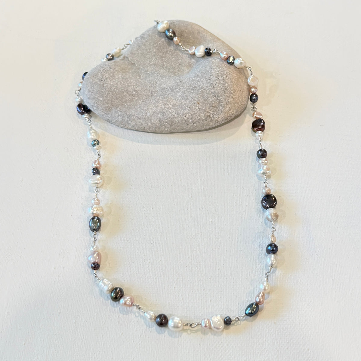 A beaded necklace with a mix of pink black, white, and iridescent pearl-like beads, draped over a smooth gray stone on a light beige background.