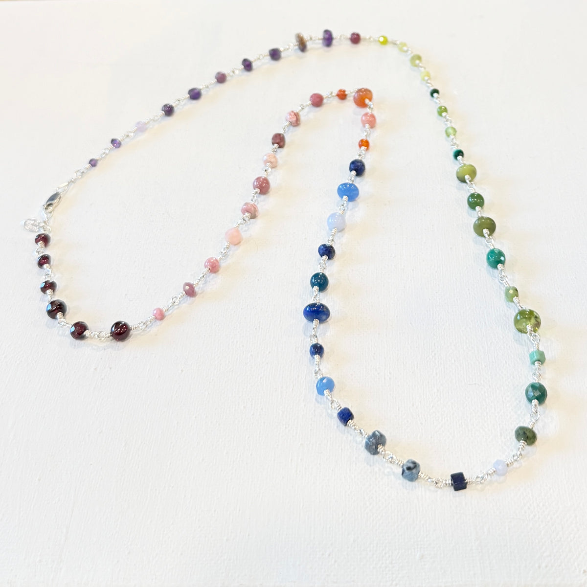 A beaded chakra necklace with multiple strands of colorful round beads in shades of purple, pink, orange, blue, green, and brown, separated by small silver spacer beads, displayed on a textured off-white background.