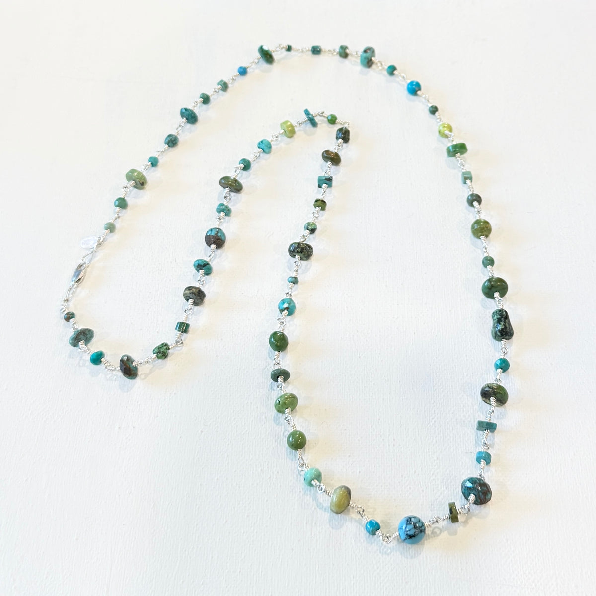 A beaded necklace with green, blue, and turquoise stones, accented by small clear crystal beads, displayed on a white textured background.
