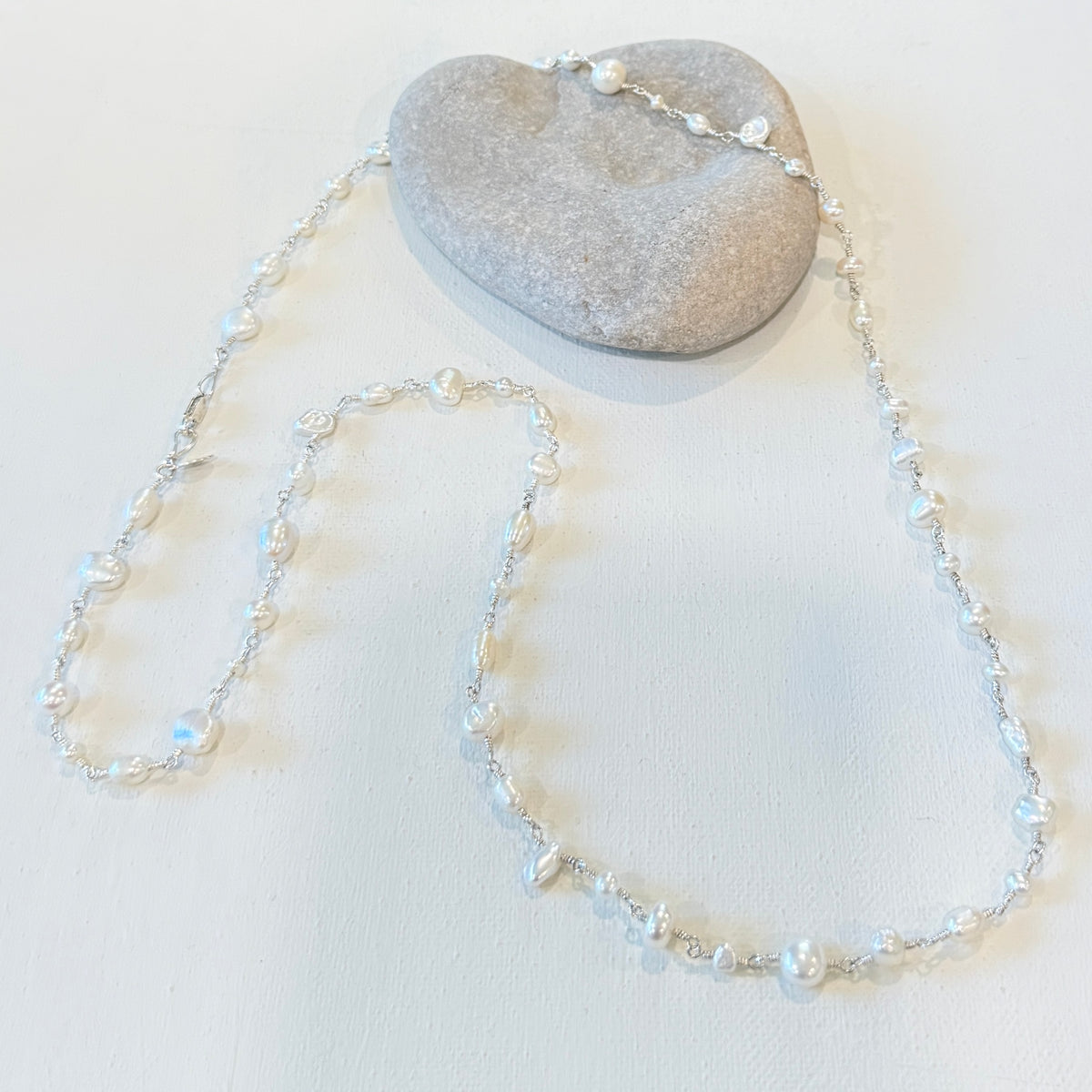 A delicate silver-toned beaded necklace with iridescent pearl beads, draped over a smooth gray stone on a light beige textured background.