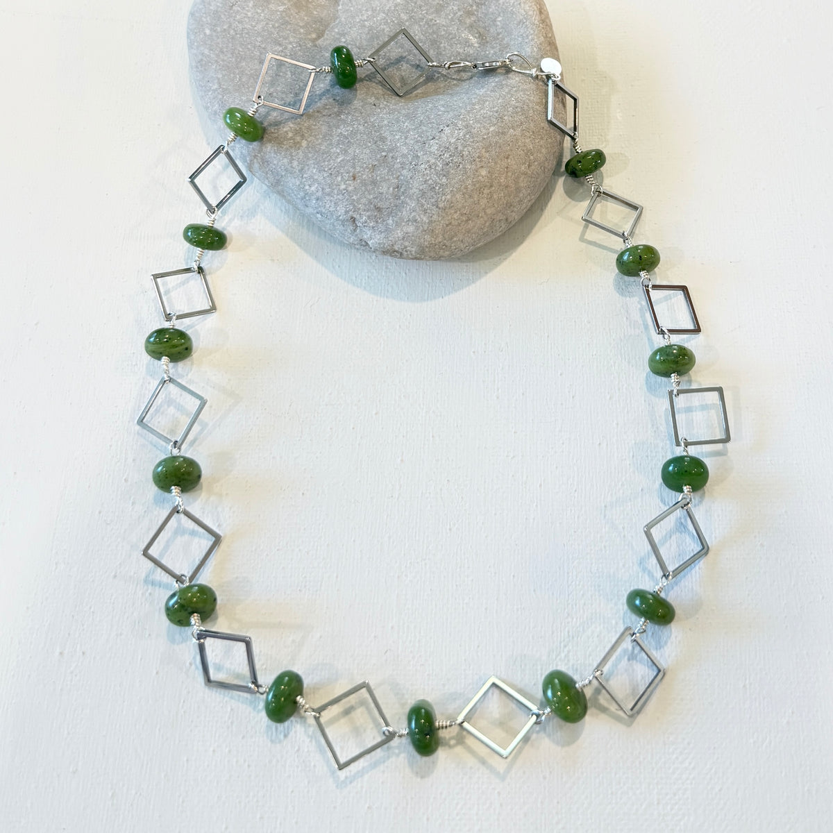 A silver-toned beaded necklace with alternating hollow square frames and oval green beads, displayed on a white surface with a gray stone prop.