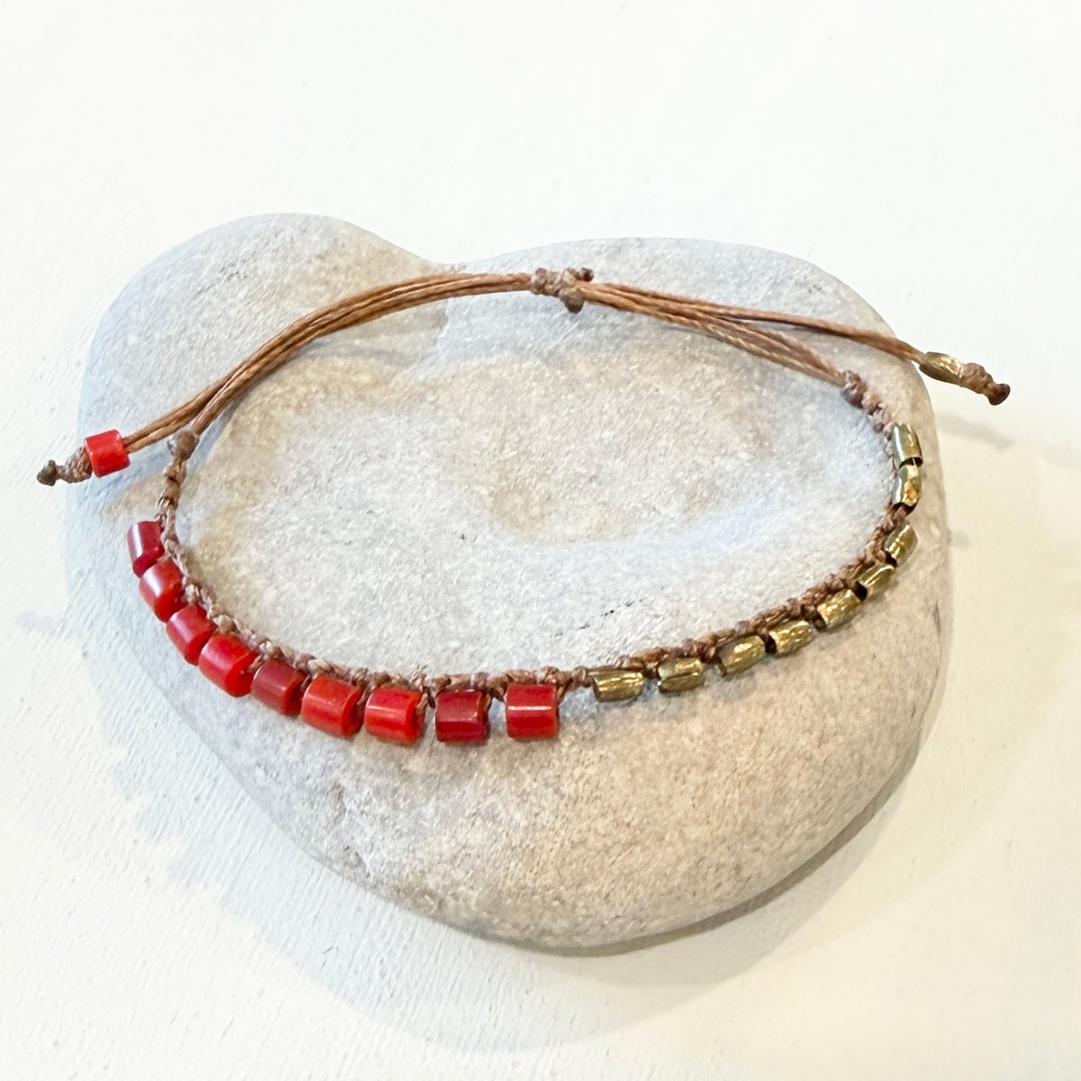 A beaded necklace with red rectangular beads and metallic gold-toned cylindrical beads on a brown cord, displayed on a light gray stone against a white background.