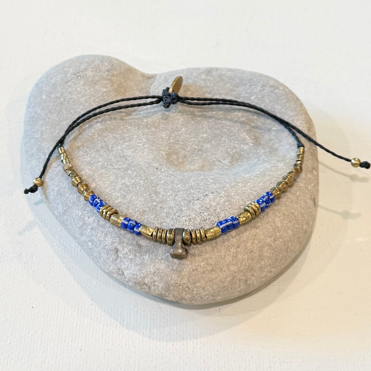 A beaded cord bracelet with blue and gold spacer beads and a small metallic charm, displayed on a light gray stone stand against a white background.