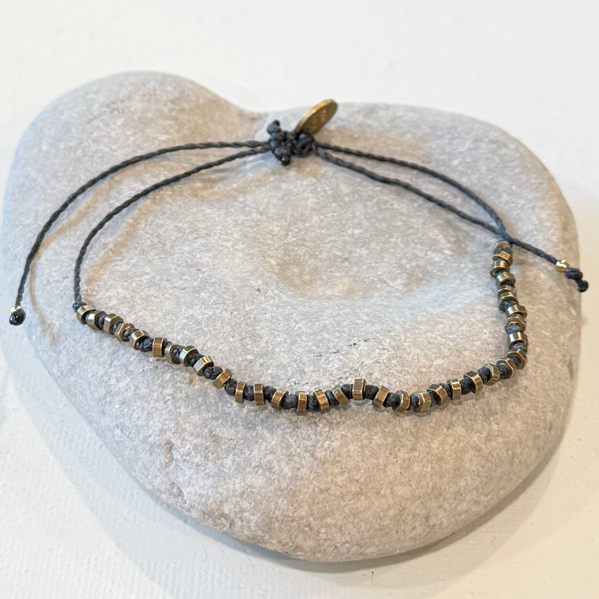 A beaded adjustable cord bracelet with metallic bronze displayed on a light gray stone stand against a white background.