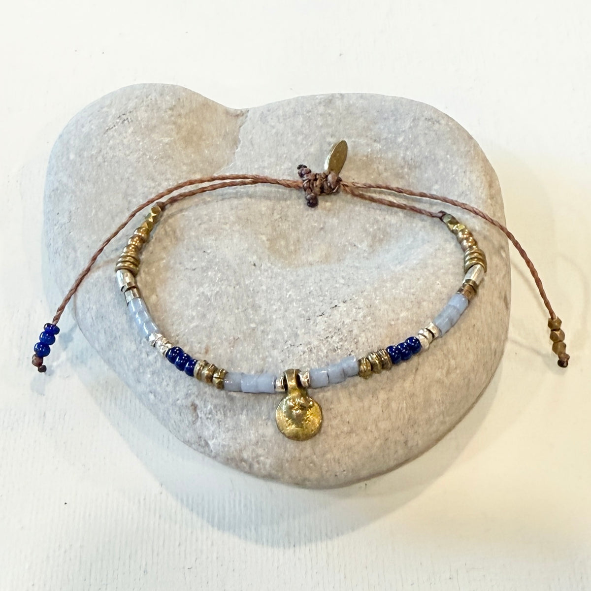 A beaded bracelet with brown cord, light blue beads, dark blue beads, gold-toned spacer beads, and a round gold charm, displayed on a heart-shaped stone stand against a white background.