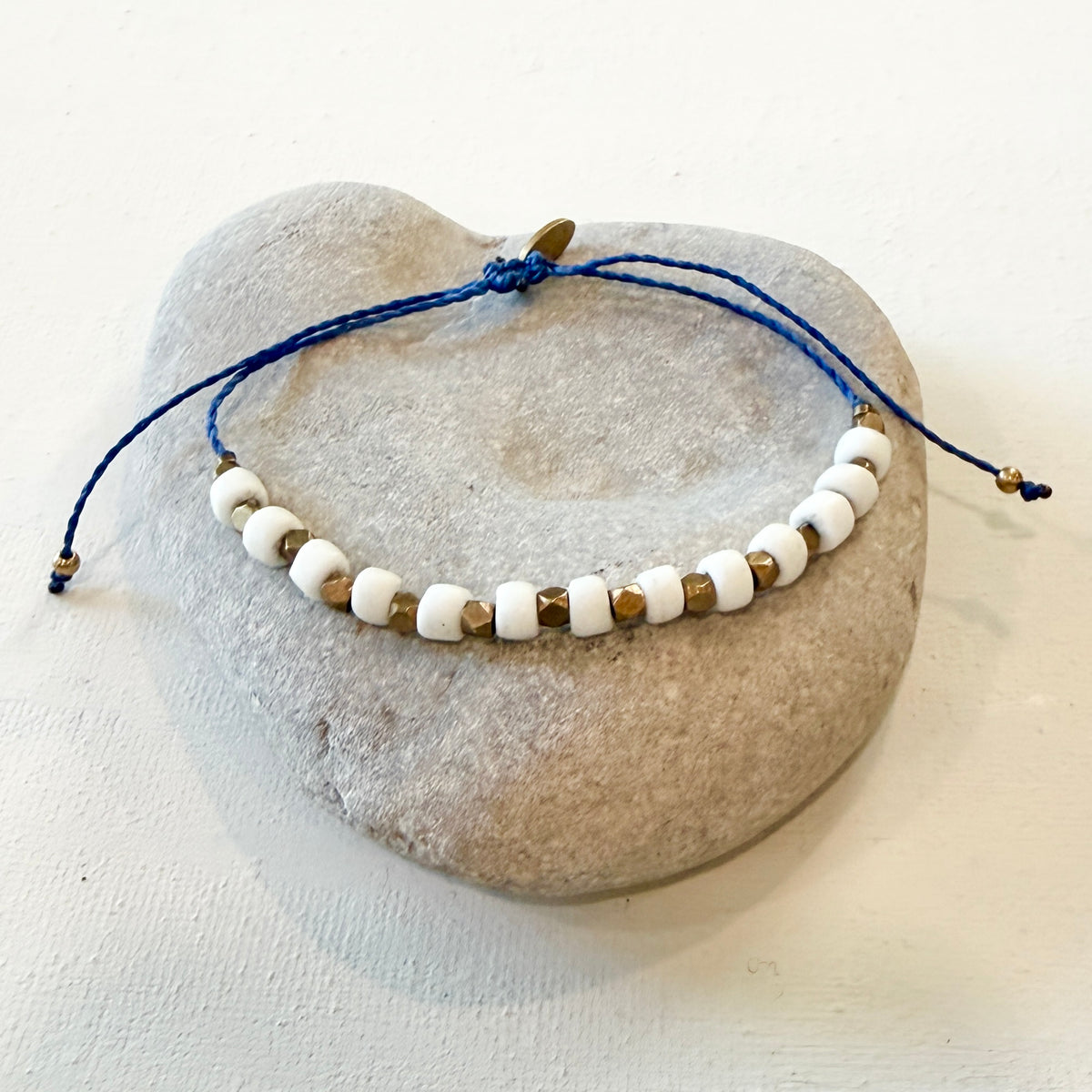 A beaded bracelet with white round beads, small metallic gold spacer beads, and a blue adjustable cord, displayed on a smooth light gray stone against a white background.