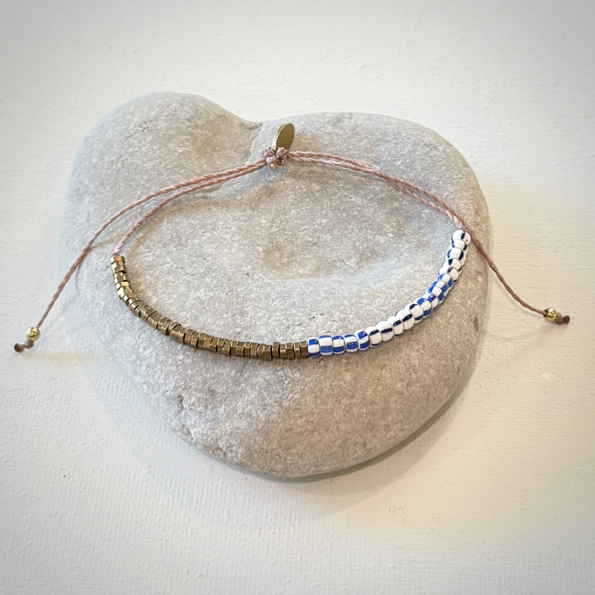 A beaded cord bracelet with gold-toned and blue-and-white patterned beads, displayed on a light gray stone stand against a white background.