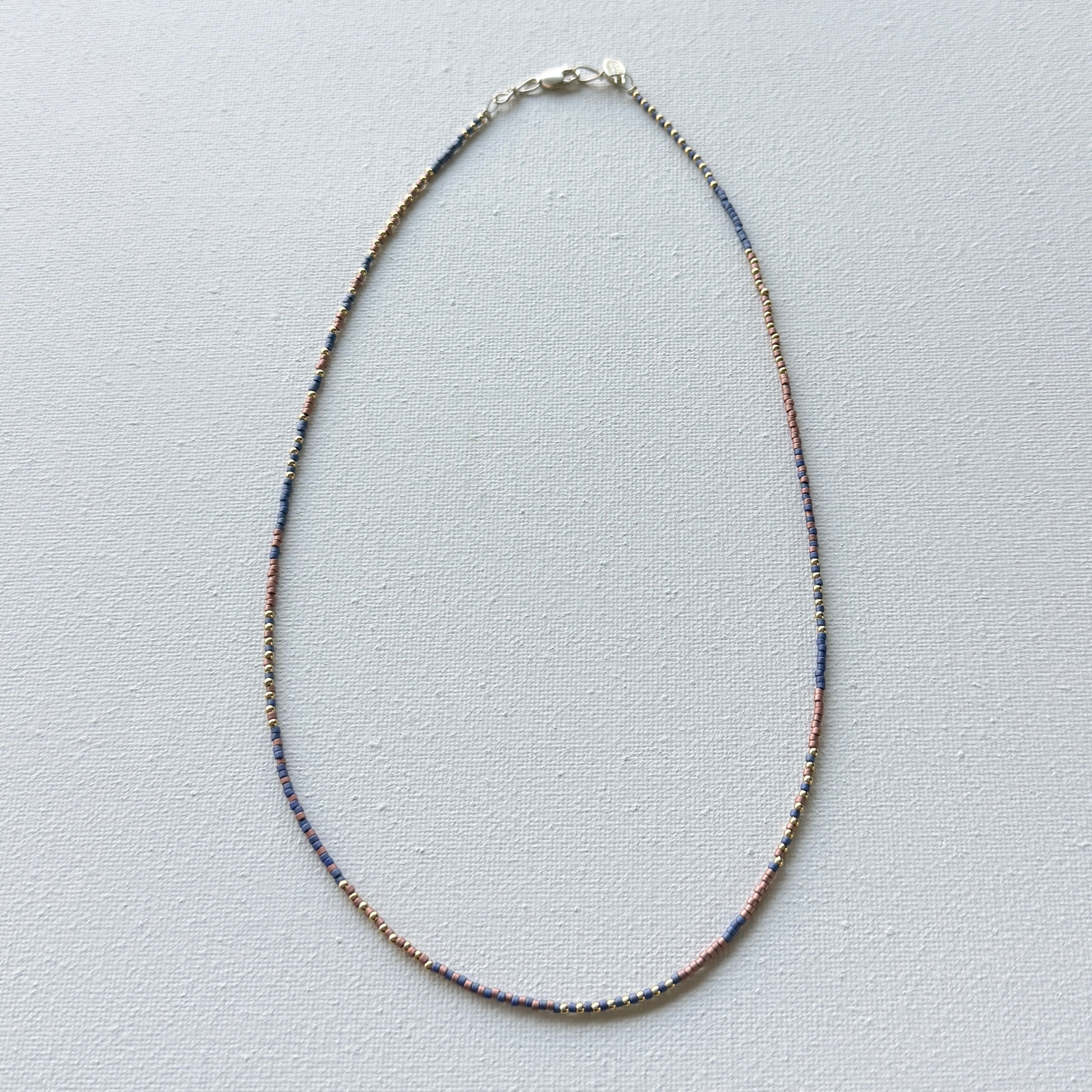 Thin beaded necklace with copper, brass and denim beads on a light gray background