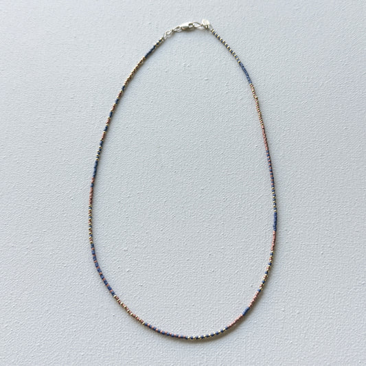 Thin beaded necklace with copper, brass and denim beads on a light gray background