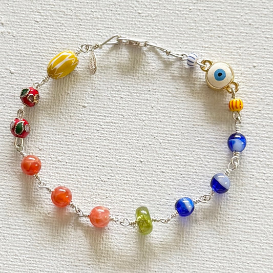 A beaded chain bracelet with a silver-tone clasp, featuring a variety of colorful glass beads including yellow, red, orange, green, blue, and a white evil eye charm, displayed on a textured white fabric background.