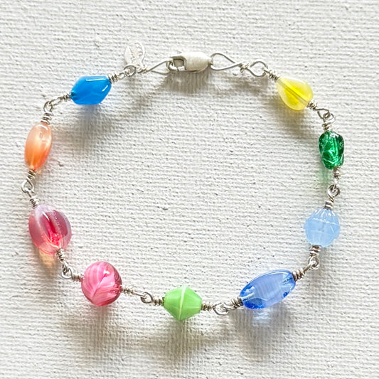 A handmade wire-wrapped beaded bracelet featuring nine colorful, faceted glass beads in shades of blue, yellow, green, pink, and light blue, arranged in a circle on a textured white fabric background.