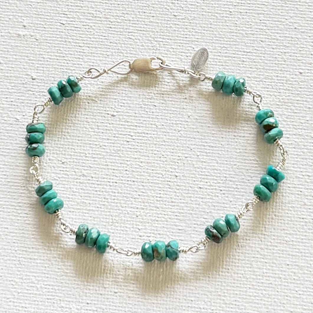 A beaded bracelet featuring small turquoise-colored stone beads hand linked on a silver wire chain with a lobster clasp, displayed on a textured off-white fabric background.