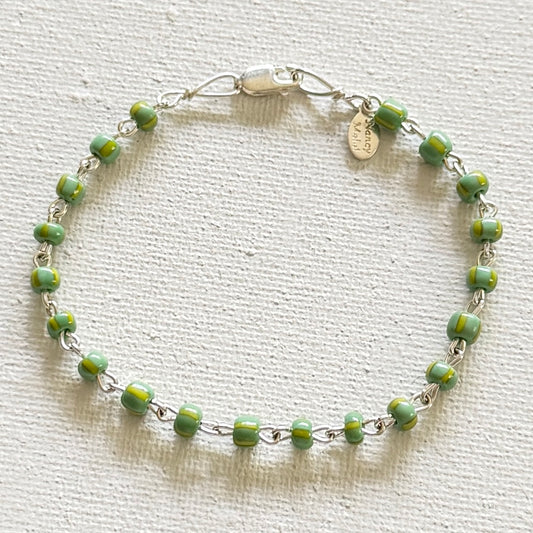 A delicate hand linked siver bracelet featuring small, round green beads with thin yellow stripes, laid out on a textured off-white fabric background.