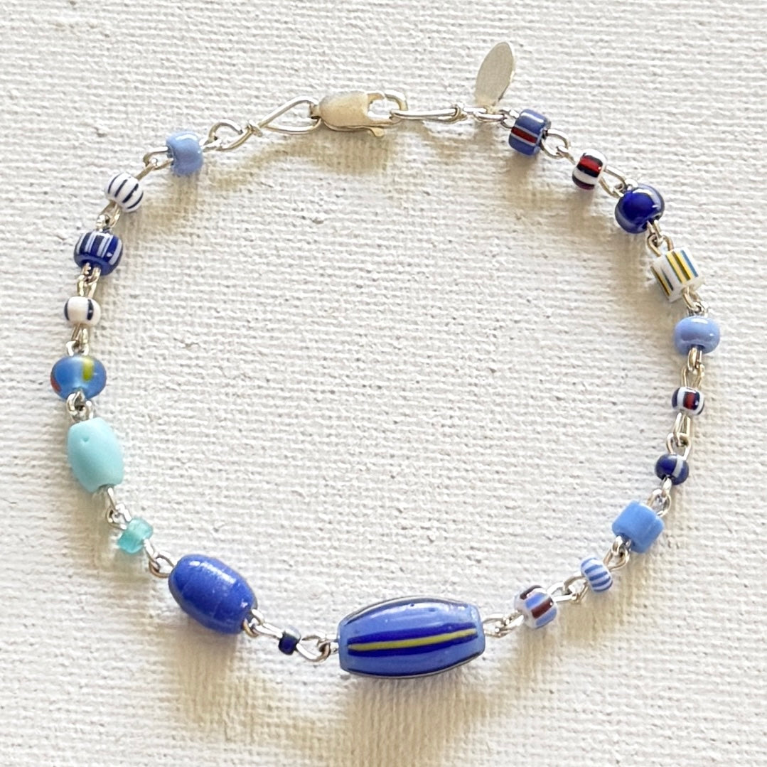 A beaded  bracelet with various blue, light blue, white, and striped glass beads, featuring a large blue striped bead at the bottom center and a silver clasp, displayed on a textured off-white fabric background.