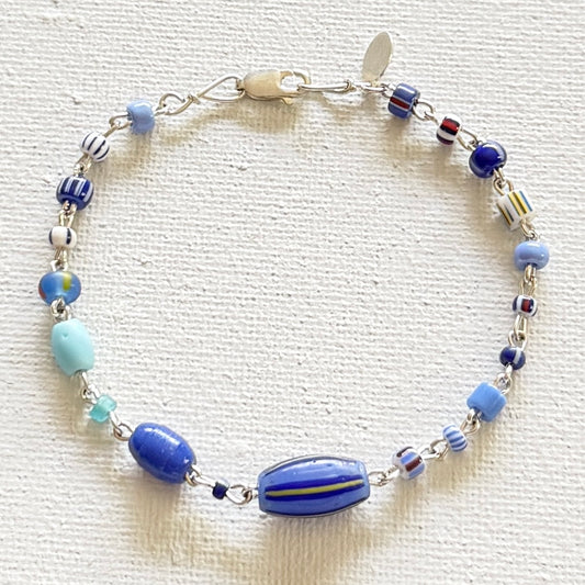 A beaded  bracelet with various blue, light blue, white, and striped glass beads, featuring a large blue striped bead at the bottom center and a silver clasp, displayed on a textured off-white fabric background.