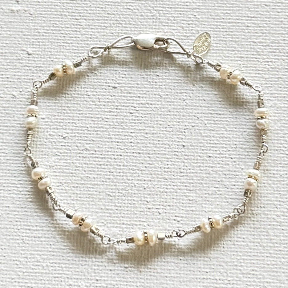 A delicate silver wire bracelet adorned with small white pearl-like beads and tiny metallic spacer beads, featuring a lobster clasp and a small round charm, displayed on a textured off-white fabric background.