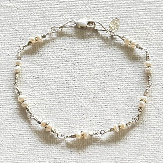 A delicate silver wire bracelet adorned with small white pearl-like beads and tiny metallic spacer beads, featuring a lobster clasp and a small round charm, displayed on a textured off-white fabric background.