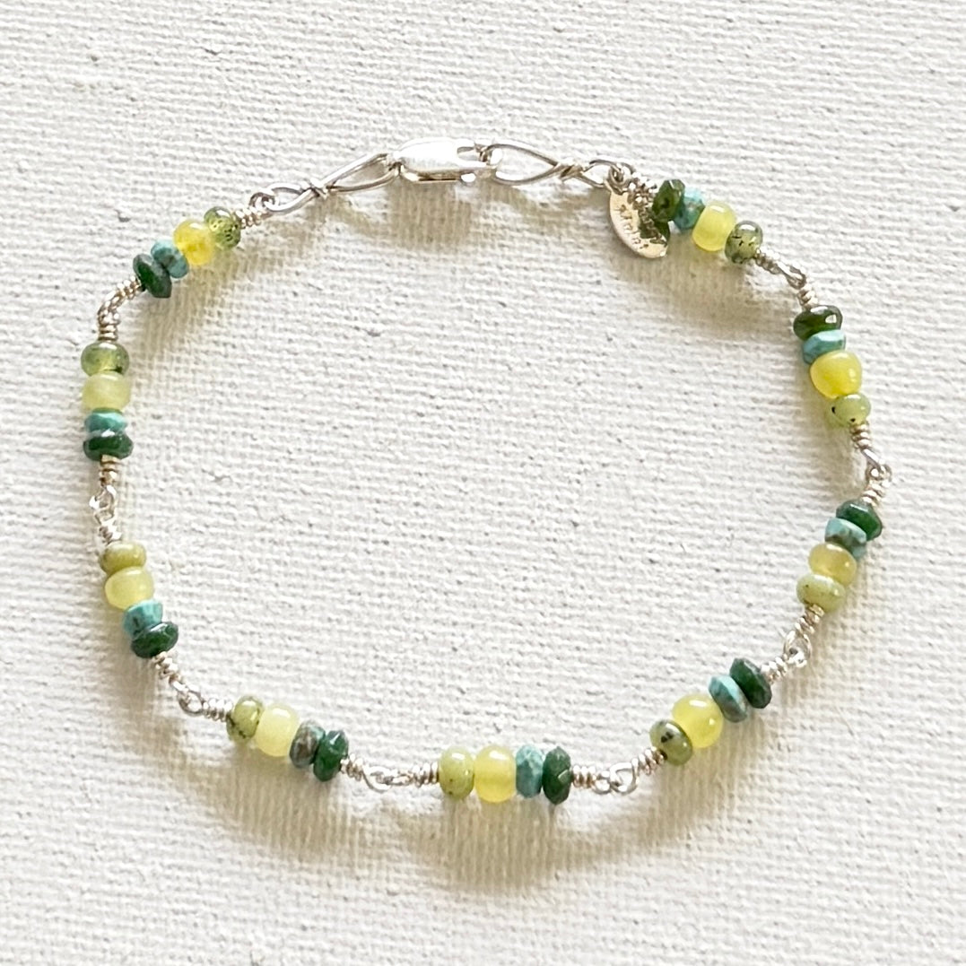 A beaded bracelet with alternating green and pale yellow marbled beads, connected by silver-toned wire spacers and a silver clasp, displayed on a textured white fabric background.