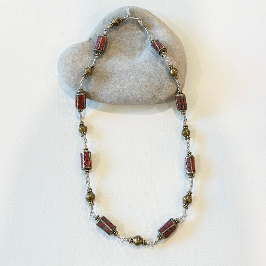 A beaded necklace with rectangular red and brown patterned beads and small metallic spacer beads, displayed draped over a smooth gray stone against a plain white wall.