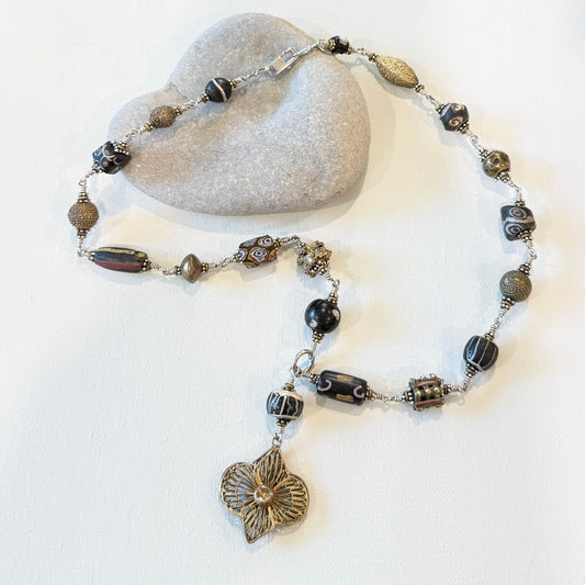 a collection with all different shapes and sized beads in black and gold with textures and lines linked with silver. A gold flower talisman on an off white background.