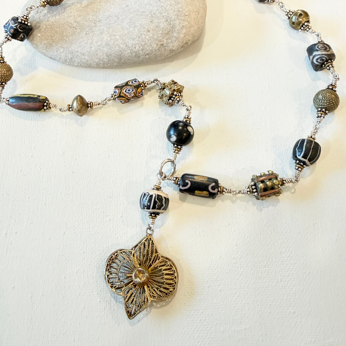 A beaded necklace with a textured bronze flower pendant, featuring black, white, and bronze glass beads on a white background with a decorative stone.