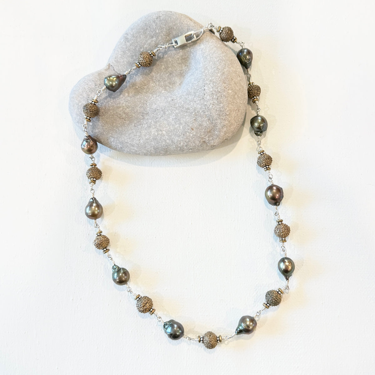A beaded necklace with dark gray iridescent pearls and textured bronze-toned spacer beads, displayed on a light gray stone prop against a white background.
