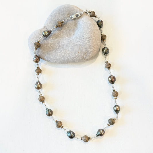 A beaded necklace with dark gray iridescent pearls and textured bronze-toned spacer beads, displayed on a light gray stone prop against a white background.