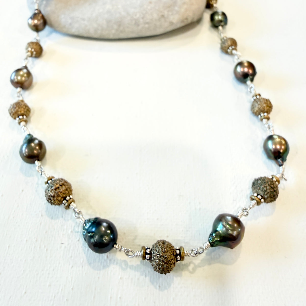 A beaded necklace featuring large, dark iridescent baroque pearls and textured brown seed beads separated by small silver spacer beads, displayed on a white surface with a neutral stone prop.