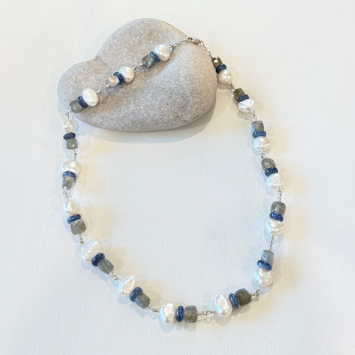 A beaded necklace and matching bracelet featuring white baroque pearls, blue faceted beads, and gray stone beads on a silver chain, displayed on a light gray heart-shaped stone against a white background.