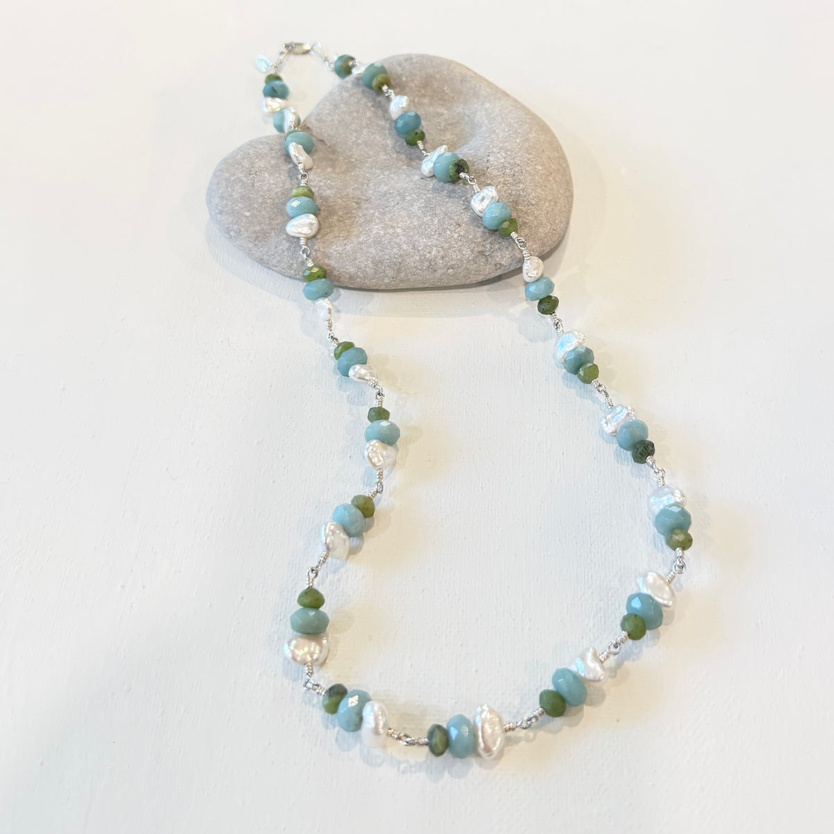 A beaded necklace with light blue, green, and white beads on a silver chain, displayed on a white surface with a smooth gray stone prop.
