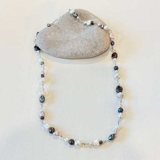 A beaded necklace with a mix of pink black, white, and iridescent pearl-like beads, draped over a smooth gray stone on a light beige background.