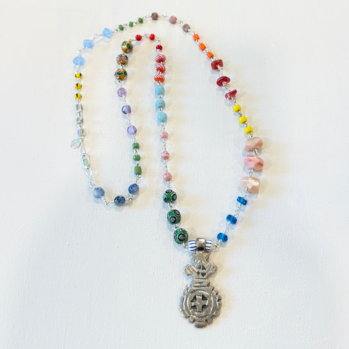 A beaded necklace with a silver-toned pendant featuring a mandala design, laid out on a white background. The necklace contains multicolored glass beads, clear faceted beads, and pink stone beads.