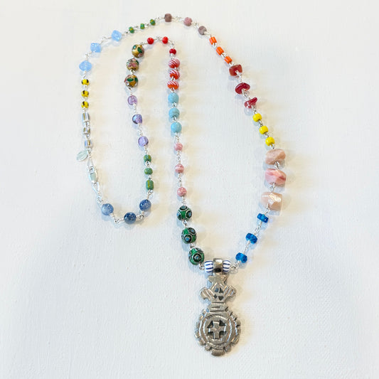 A beaded necklace with a silver-toned pendant featuring a mandala design, laid out on a white background. The necklace contains multicolored glass beads, clear faceted beads, and pink stone beads.