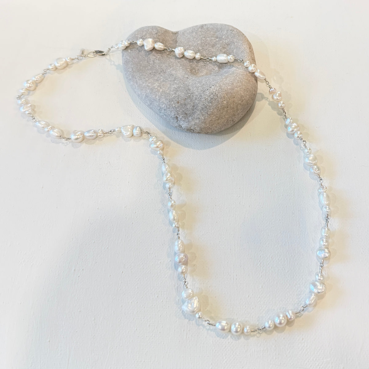 A delicate necklace made of small white freshwater pearls on a silver chain, draped over a smooth gray stone against a plain off-white background.