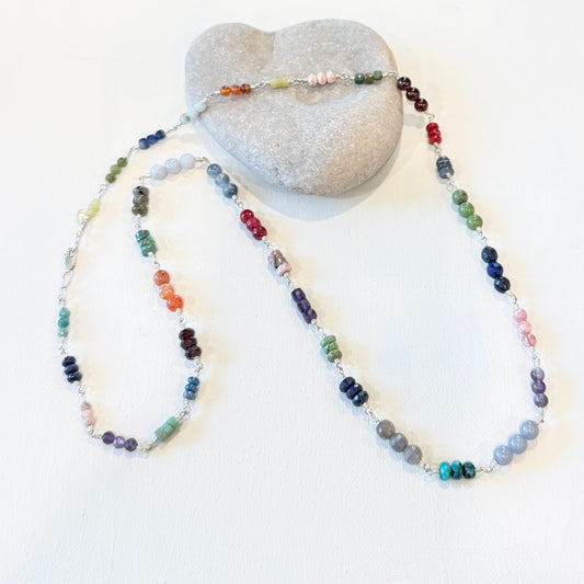 A long beaded necklace with multicolored gemstone beads and hand linked silver draped around a smooth gray stone on a white background.