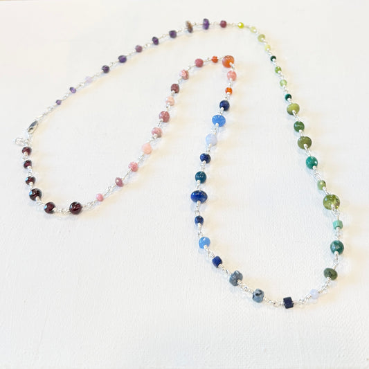 A beaded chakra necklace with multiple strands of colorful round beads in shades of purple, pink, orange, blue, green, and brown, separated by small silver spacer beads, displayed on a textured off-white background.