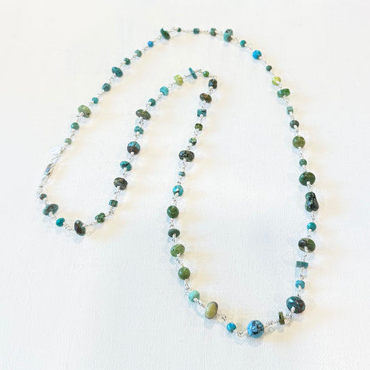 A beaded necklace with green, blue, and turquoise stones, accented by small clear crystal beads, displayed on a white textured background.