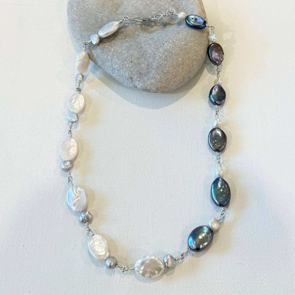 A beaded necklace featuring iridescent baroque pearls, dark gray pearls and hand linked silver  displayed on a light gray stone against a white background.