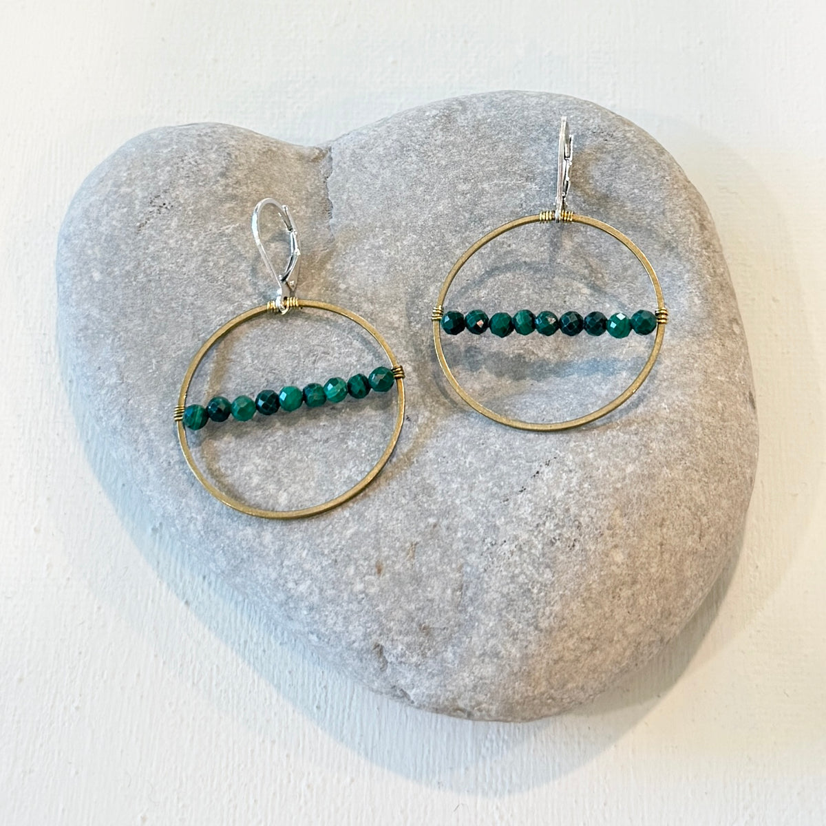 A pair of gold and silver hoop earrings with green beaded horizontal bars, displayed on a light gray heart-shaped stone against a white background.