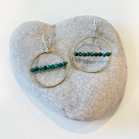 A pair of gold and silver hoop earrings with green beaded horizontal bars, displayed on a light gray heart-shaped stone against a white background.