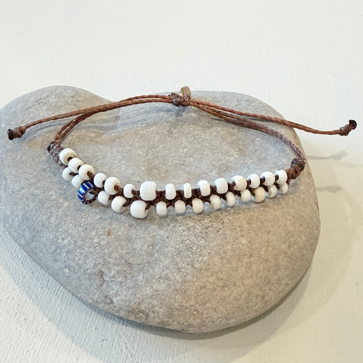 A double-strand beaded bracelet with white round beads, brown cord, and a small blue and white evil eye charm, displayed on a smooth gray stone against a light off-white background.