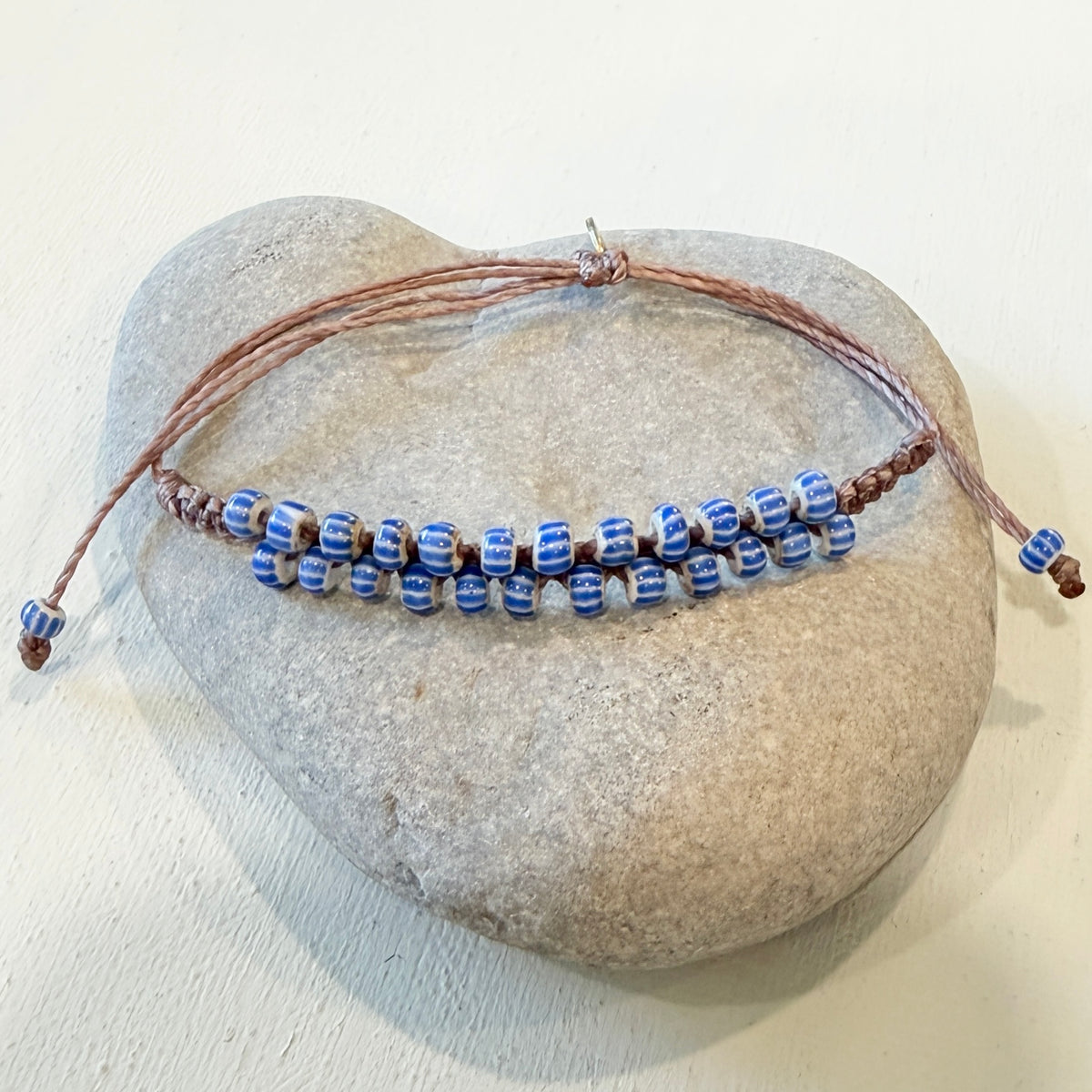 A handmade beaded bracelet with blue and white striped glass beads on a brown cord, displayed on a smooth gray stone against a light beige background.
