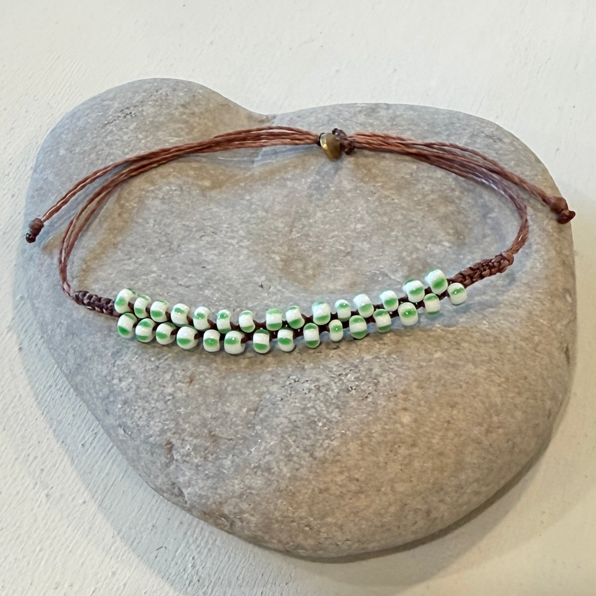 A beaded bracelet with brown cord and small white beads with green and black stripes, displayed on a smooth gray stone against a light beige background.