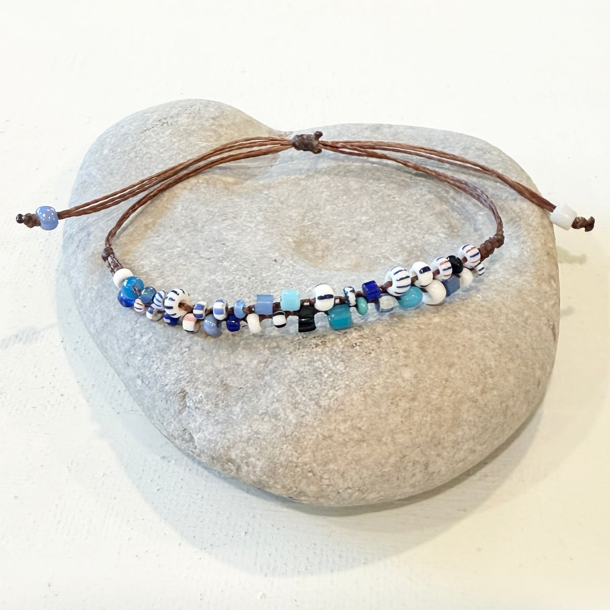 A beaded adjustable cord bracelet displayed on a smooth light gray stone. The bracelet features brown cords, small white beads with blue and black patterns, and translucent blue beads.
