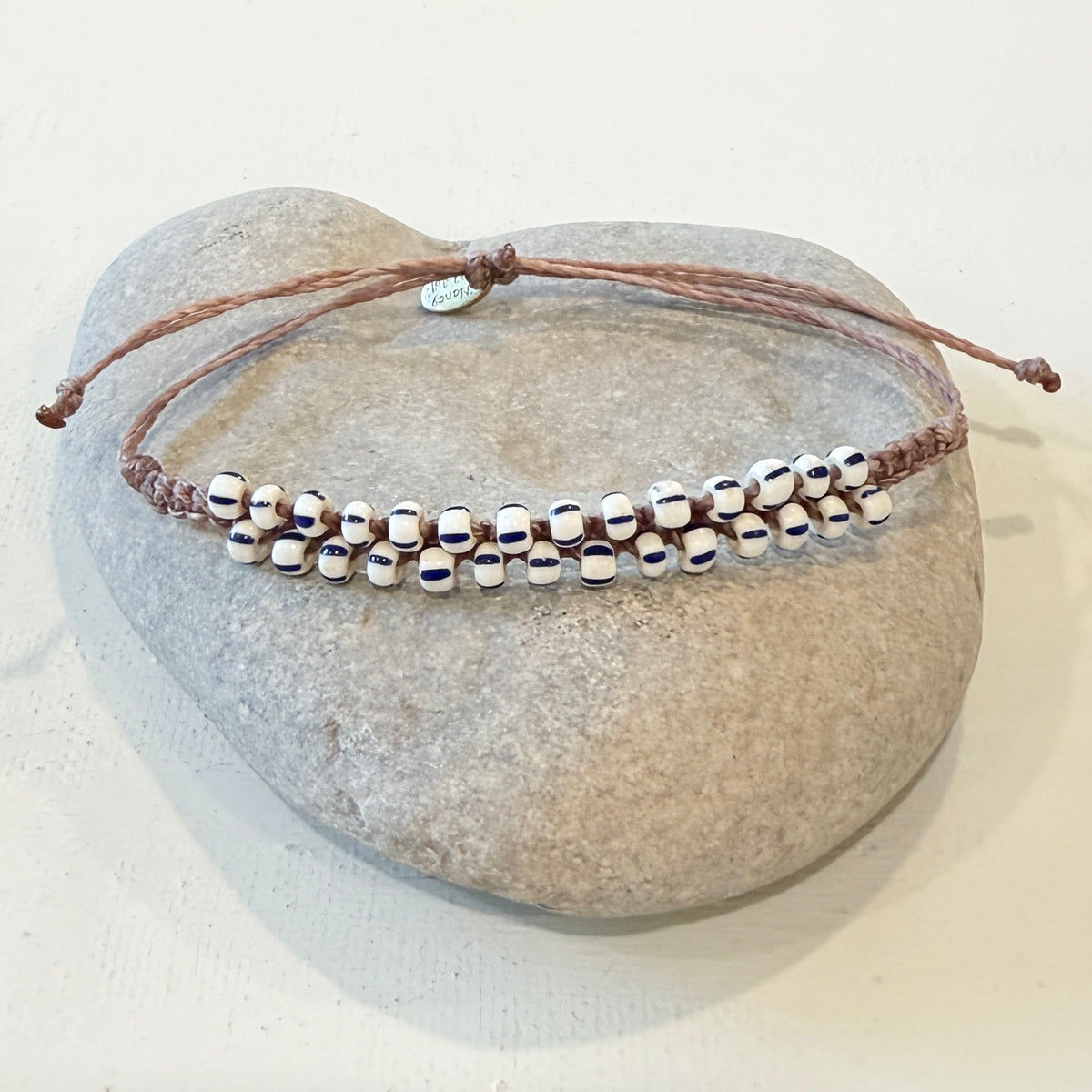 A beaded adjustable cord bracelet displayed on a smooth gray stone against a light neutral background. The bracelet features a brown cord and a row of small white beads with dark blue centers.